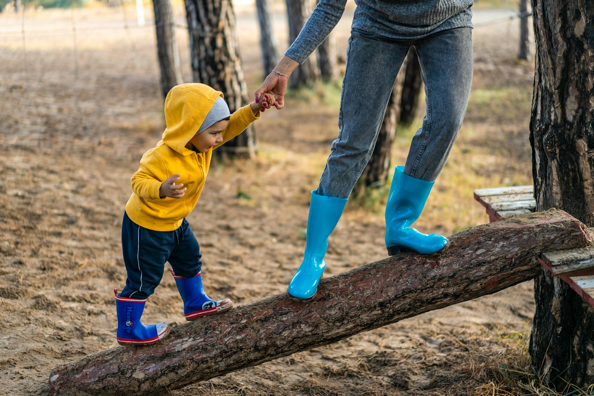 Un enfant avec des bottes en caoutchouc bleu et rouge et un sweat à capuche marche sur un tronc d'arbre couché avec l'aide d'un adulte. S'engager, se mettre en mouvement.